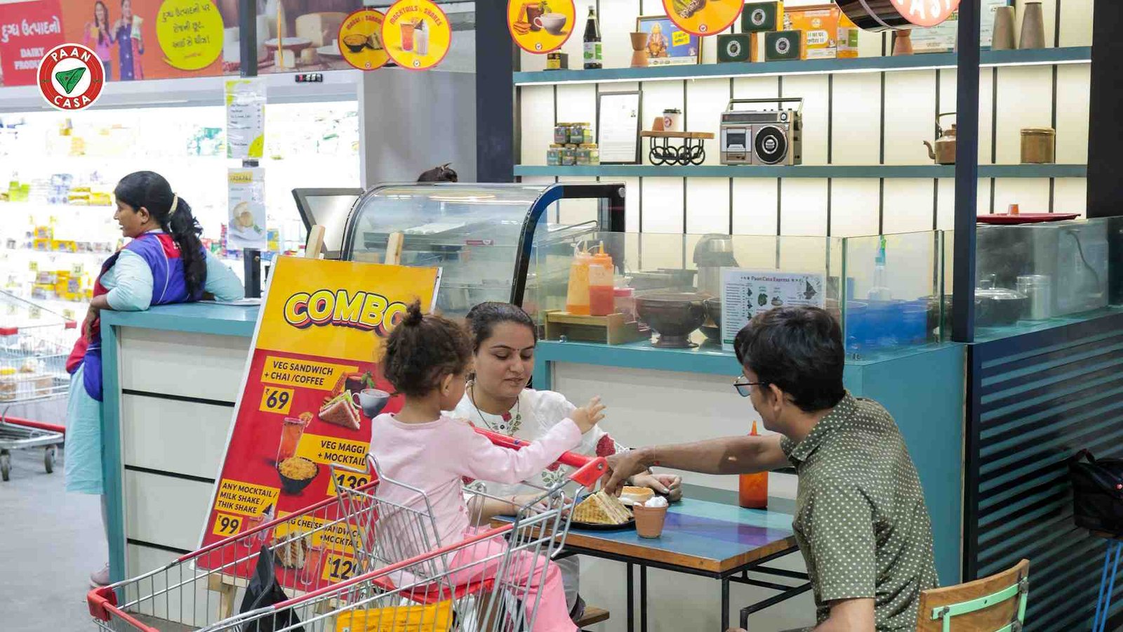 Family enjoying sandwiches at a Paan Casa café inside a supermarket with a child in a shopping trolley.